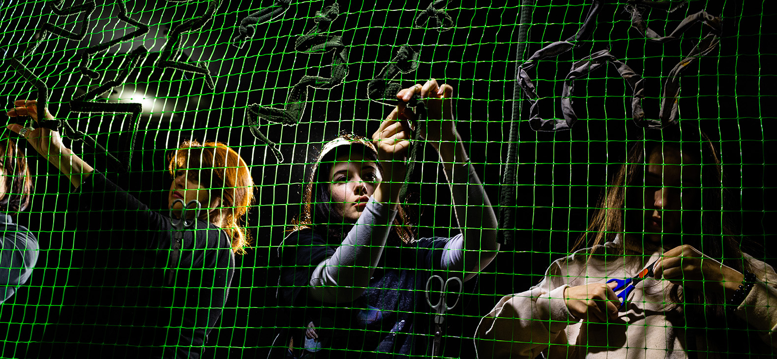 The Thin Line - Exhibition of Ukrainian photographs; Image: Volunteers knotting camouflage nets, young women in the spotlight behind a green net; Photographer: Kateryna Moskalyuk 