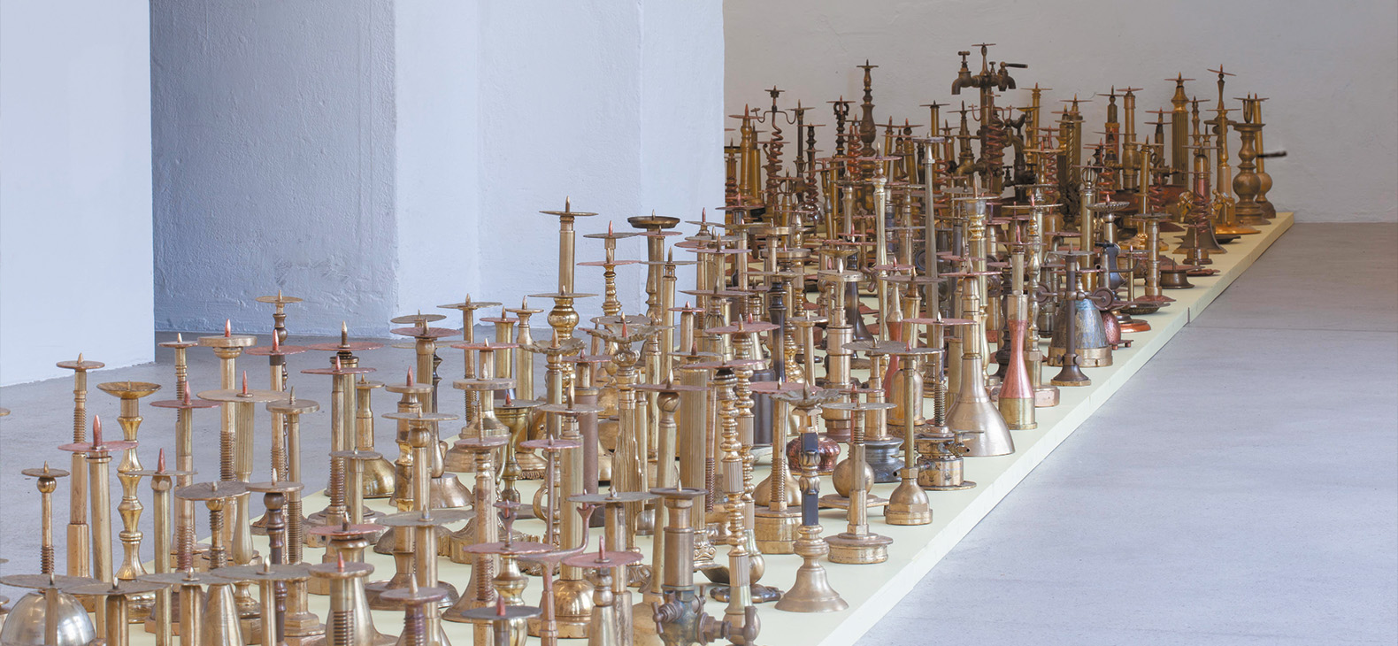 Rebekka Bauer: As found as objects, exhibition view Kunstraum Ortloff Leipzig, 2019; photo: Rebekka Bauer; photograph of an installation of 550 shiny metal objects (mostly candlesticks) on a limited track on the floor of a white room.